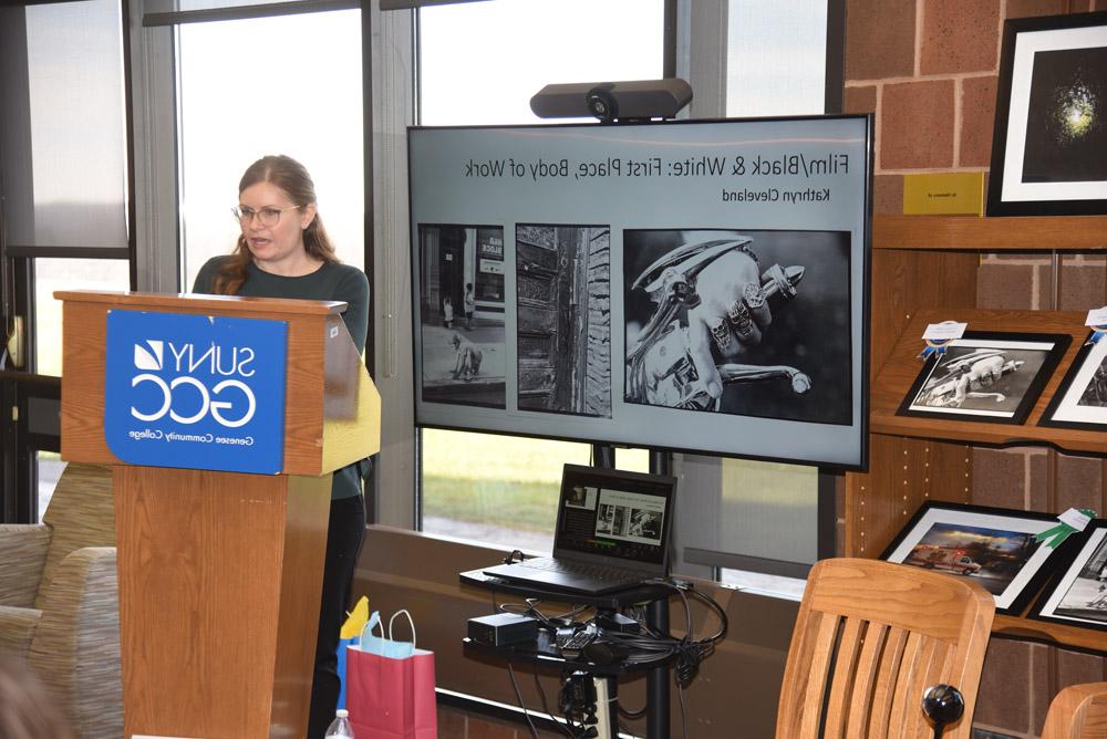 系统 & Electronic Services Librarian Liz Simmons speaking at ceremony, slides showing Film/B&W First Place winning photos behind